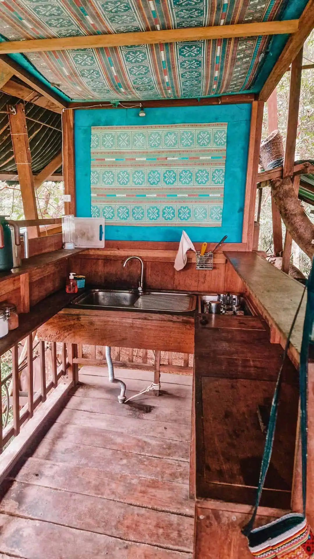 A rustic treehouse kitchen with wooden countertops and a small stainless steel sink. The walls are decorated with blue patterned fabric, and the ceiling has a colorful woven design. Shelves hold jars, metal kettles, and cooking essentials, while natural light filters through the open-air structure in Laos.
