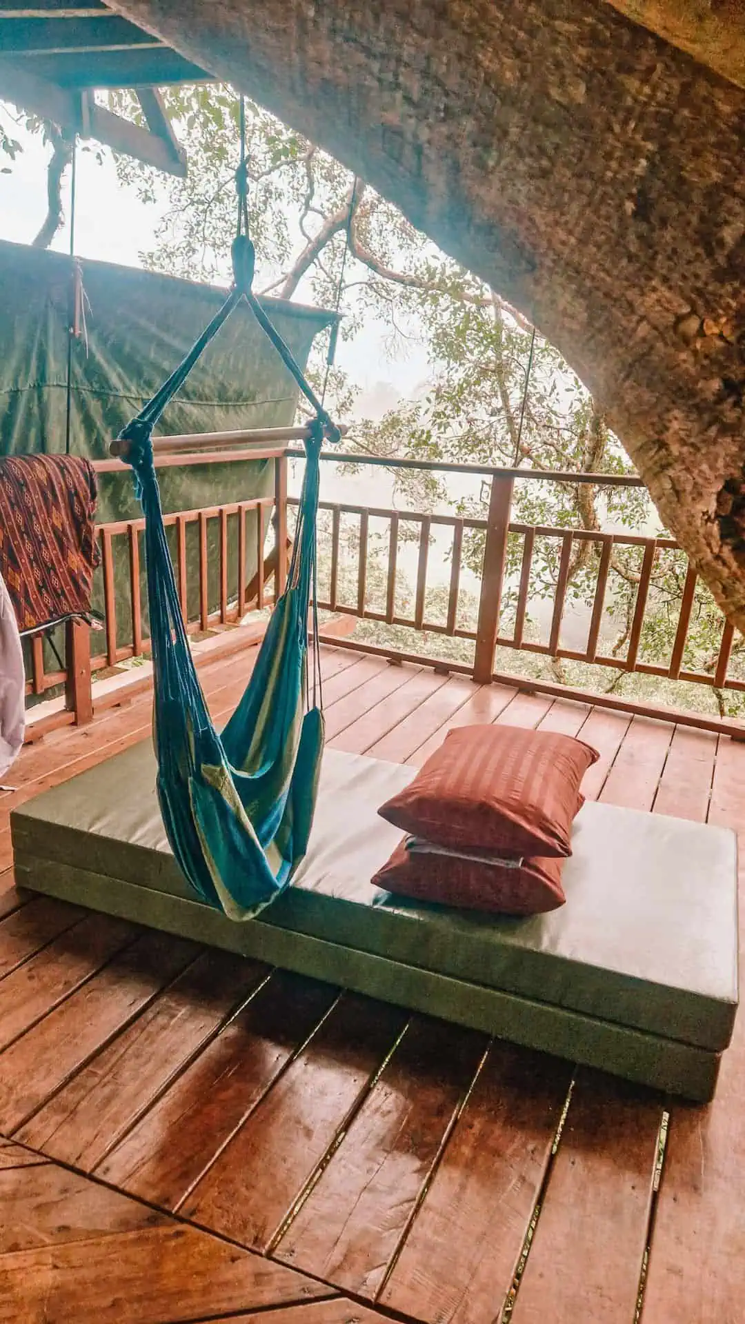 A cozy treehouse balcony with a floor mattress, two pillows, and a hanging blue hammock chair. The wooden deck is surrounded by a railing, and a green tarp hangs in the background. A large tree trunk extends into the space, blending nature with the rustic design in Laos.