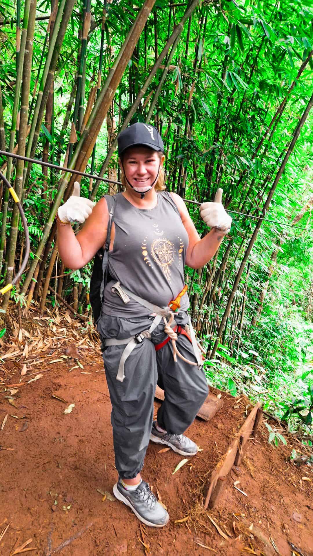 A woman dressed in outdoor adventure gear stands on a dirt path surrounded by tall bamboo. She wears a harness, gloves, and a helmet, giving a thumbs-up and smiling at the camera. A zipline cable is attached nearby, indicating a jungle adventure experience.