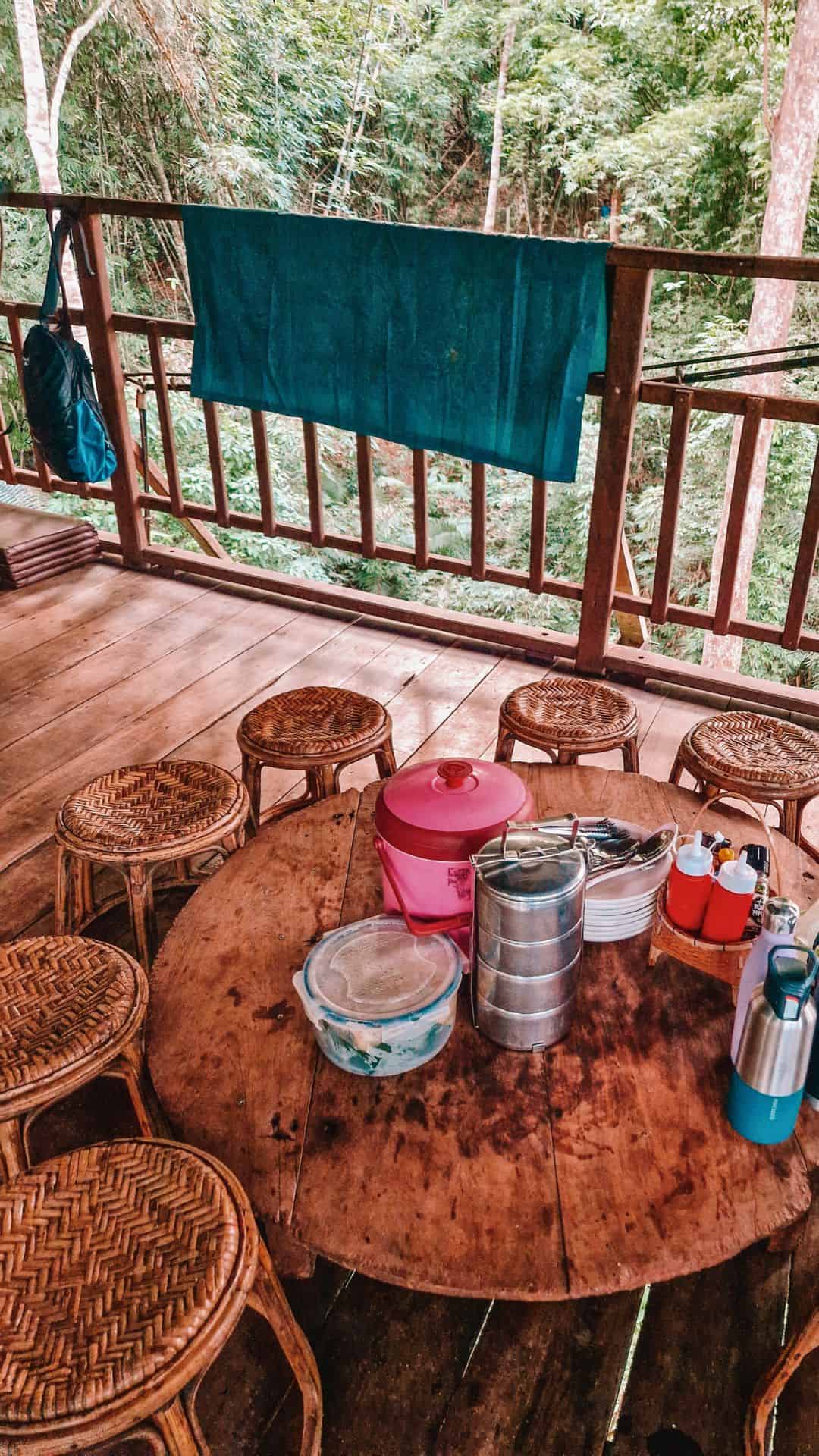 A rustic wooden dining table with woven rattan stools, set with food containers, water bottles, and condiments, on a treehouse balcony overlooking the jungle.