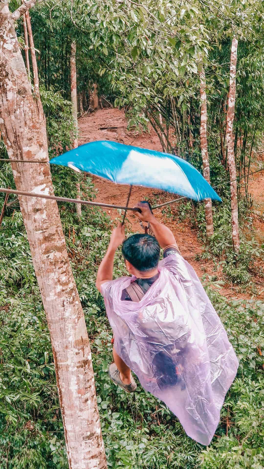 A person mid-zipline rides over a dense jungle, holding a blue umbrella. They wear a translucent rain poncho and casual outdoor attire. The zipline extends between trees, with the forest floor visible far below in Laos.